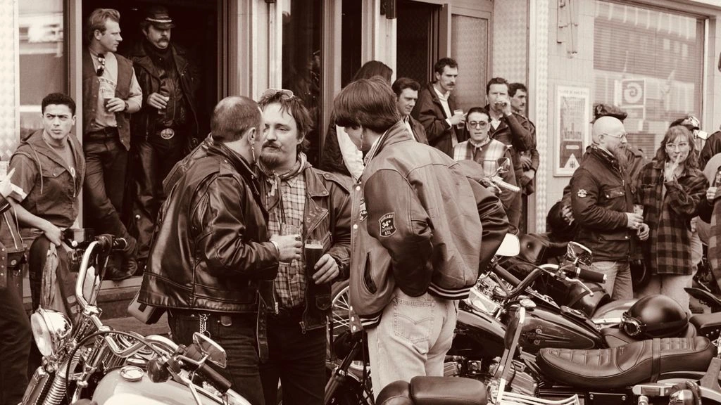 Riders gathered around a roadside table at golden hour, helmets off, two motorcycles parked in the background.